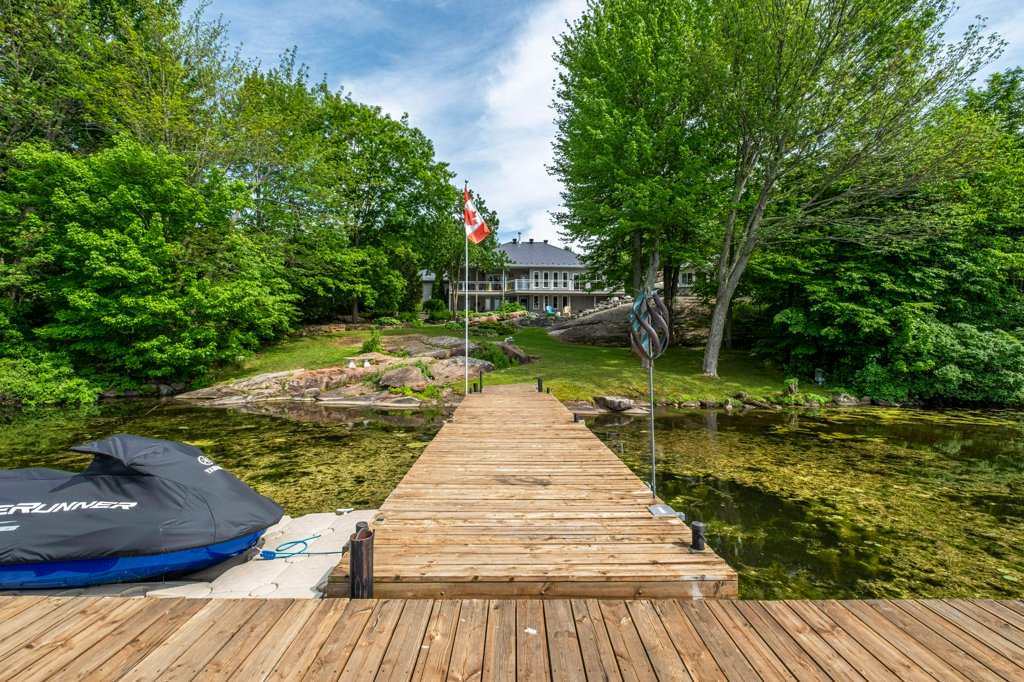 Waterfront estate in the Kingston area seen from the end of a private dock, with a Canadian flag, mature trees and a sweeping lawn leading up to the home